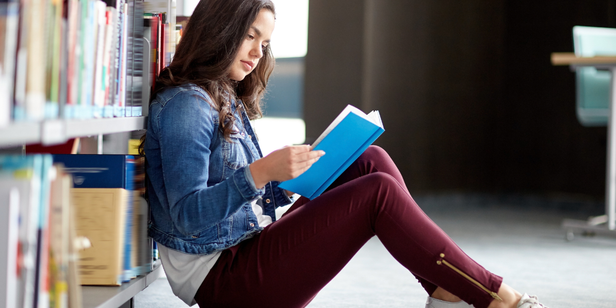 A young woman in denim jacket sits on library floor reading a blue book, surrounded by colorful bookshelves, showcasing the focused concentration that comes from the benefits of slow reading