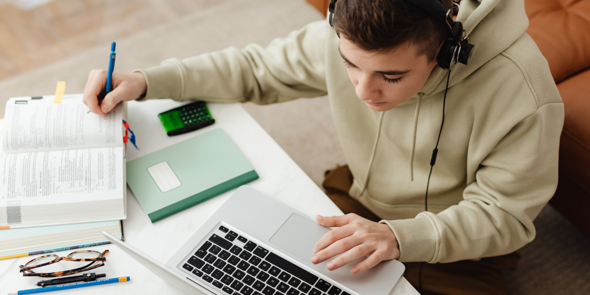 Teen learning to write news copy on laptop for student newspaper with headphones and study materials