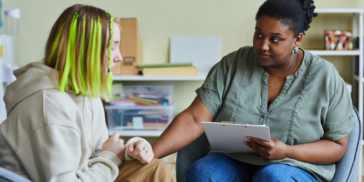 Female counselor or therapist having supportive conversation with young child in comfortable office setting with educational materials and toys in background