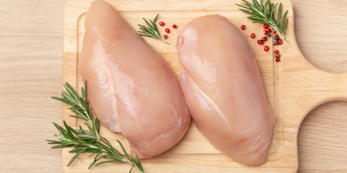 Two boneless skinless chicken breast fillets on wooden cutting board garnished with fresh rosemary and pink peppercorns, showing white meat composition for nutrition education