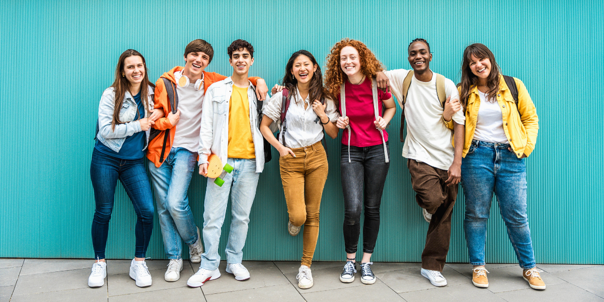 Seven diverse teenagers standing together with arms around each other against turquoise wall, showing healthy peer relationships and social bonding skills