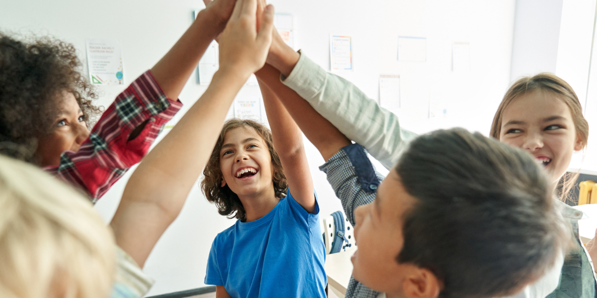 Group of teenagers putting hands together for team high-five in bright classroom setting, illustrating cooperation and group bonding behaviors