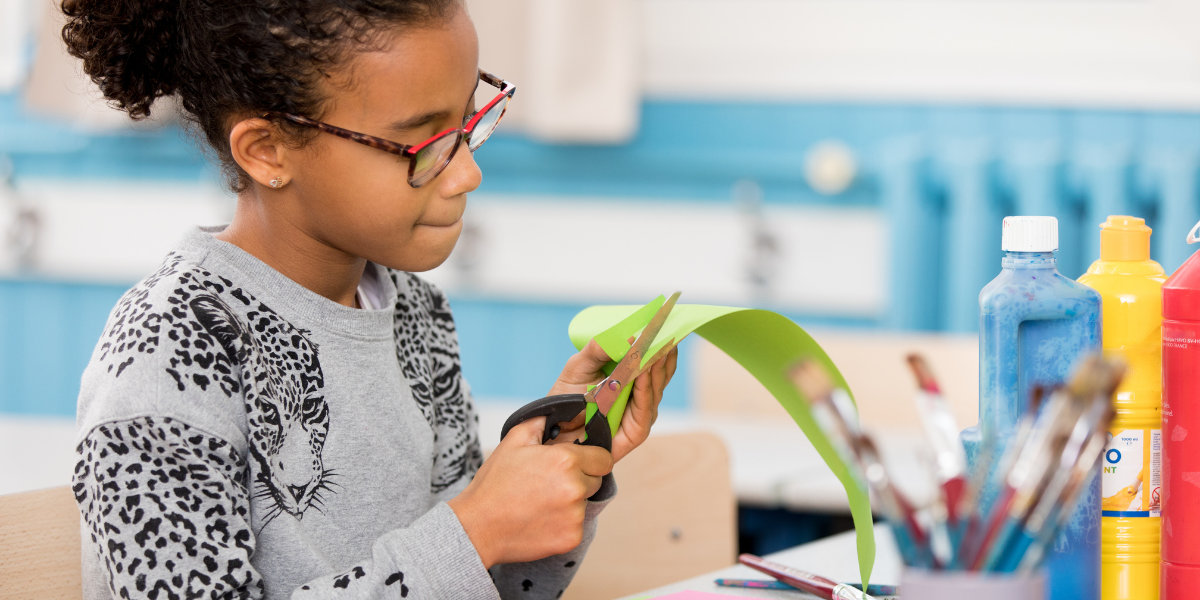 A focused young girl with glasses carefully cuts green paper during an art class, exemplifying the concentration and fine motor skills developed when schools teach art in schools as part of comprehensive education.