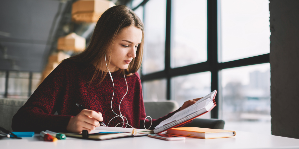 A blonde woman in red sweater sits at a table with multiple open books and headphones, taking notes while reading - illustrating how the benefits of slow reading enhance learning and comprehension