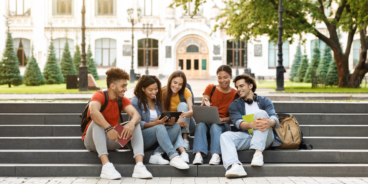 Five diverse students sitting together on campus steps sharing devices and studying collaboratively, demonstrating healthy peer relationships and cooperative problem-solving skills