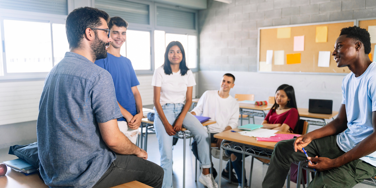 Male teacher leading engaged classroom discussion with diverse group of students sitting in circle, demonstrating collaborative learning and open communication techniques
