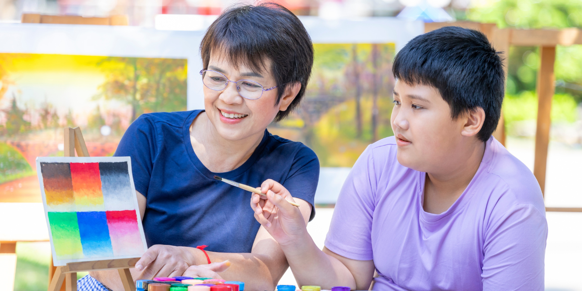 A smiling teacher guides a young student through a painting lesson outdoors, demonstrating the personal mentorship approach used to teach art in schools and foster creative development.