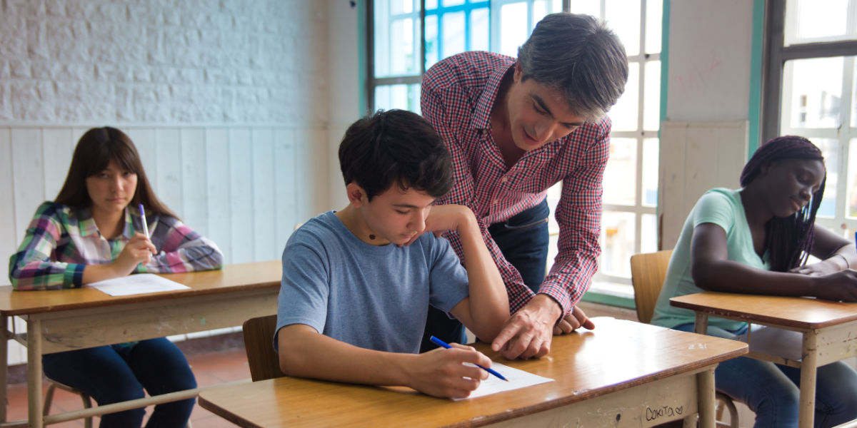 Male teacher in checkered shirt helping teenage student with schoolwork in classroom while other students work independently at their desks