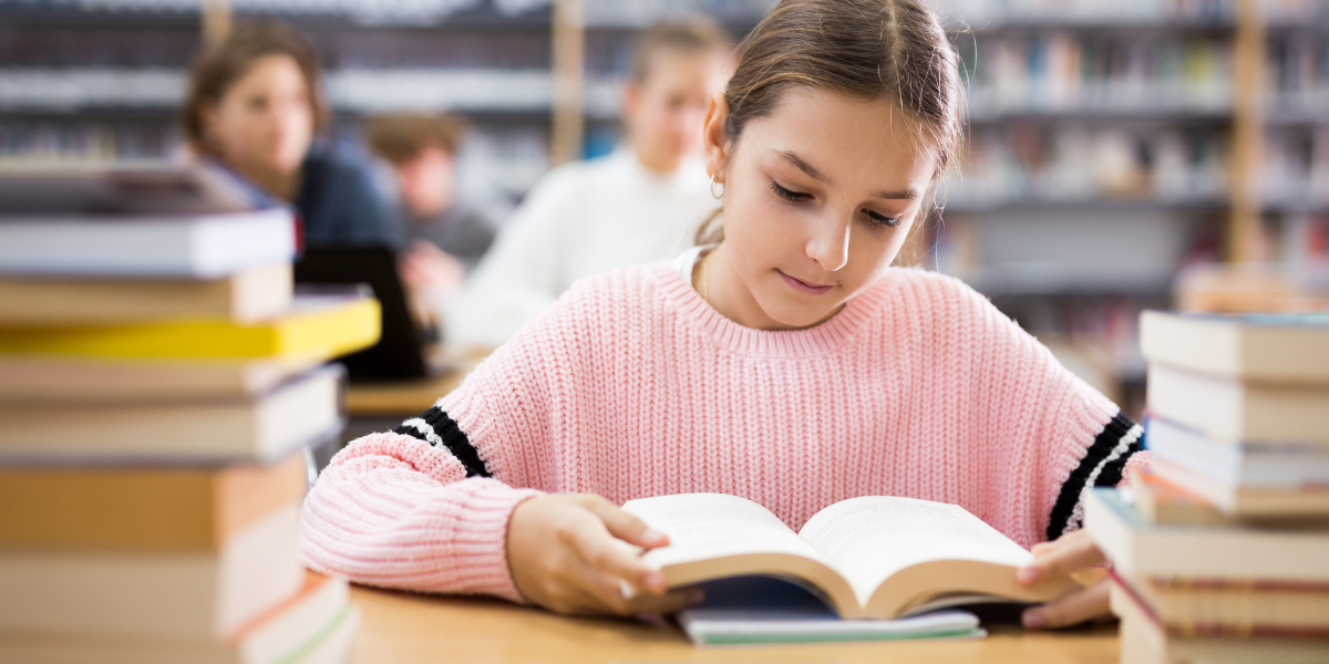 Concentrated teenage girl in pink sweater reads textbook in quiet library setting, illustrating how proper study environments help teens concentrate for longer periods during academic tasks.