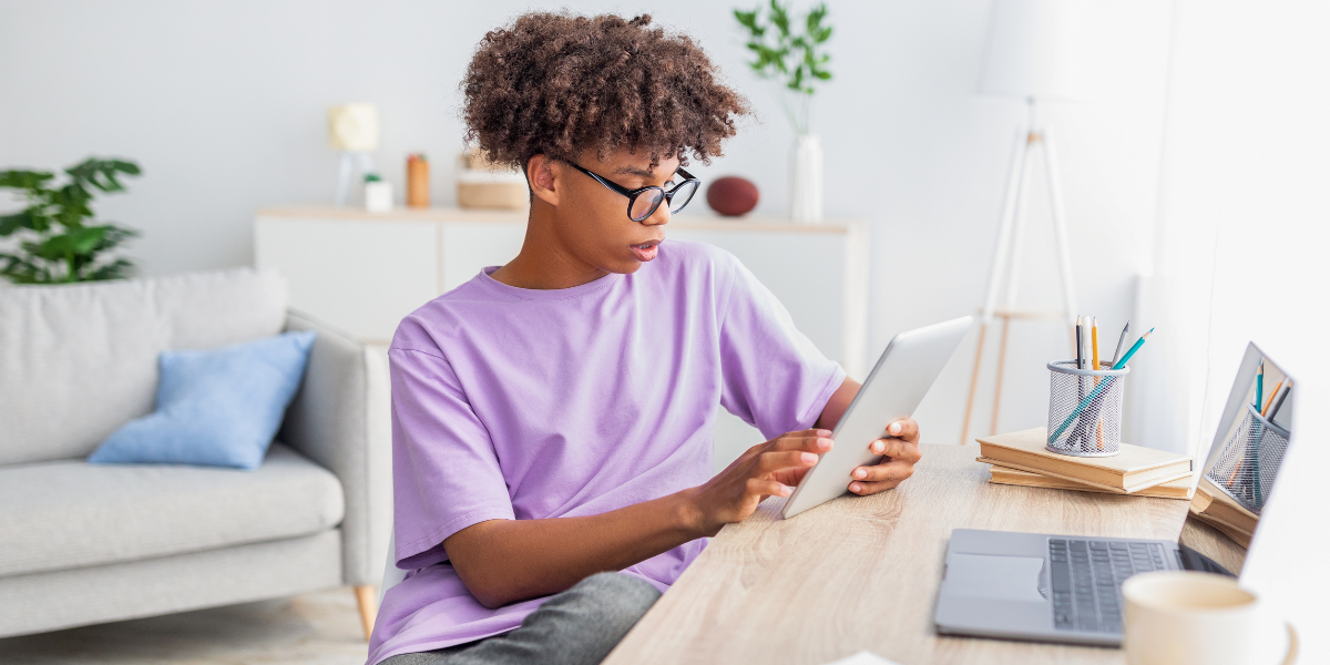 Focused teenager wearing glasses uses tablet for studying at organized desk with laptop and books, demonstrating effective ways to help teens concentrate in a distraction-free environment.