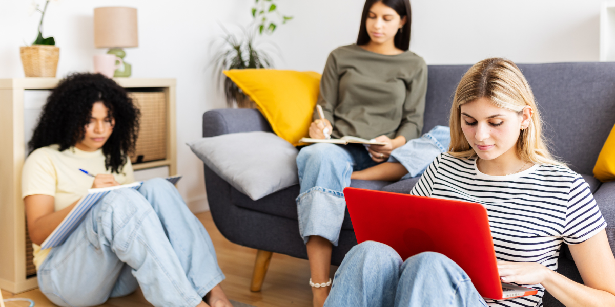 Three diverse women engage in quiet reading activities in a contemporary living space with grey couch and yellow accent pillow, each focused on their individual books and materials in a peaceful group study environment