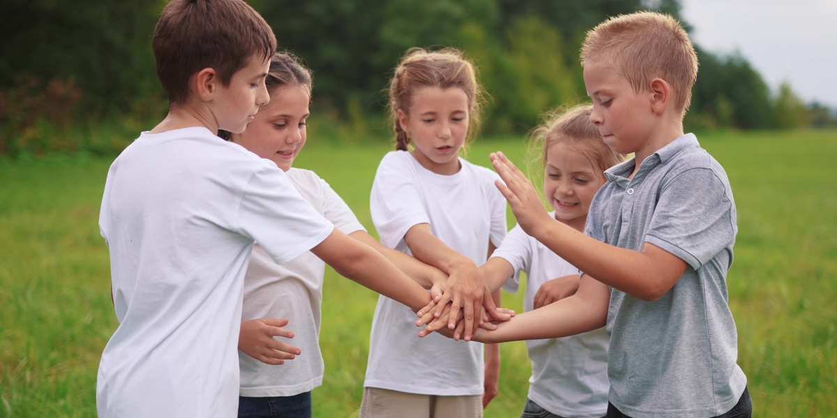 Five children stacking hands together in outdoor setting, practicing collaboration and team-building skills that develop into teenage social competence