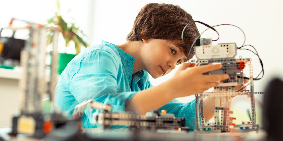 Focused boy in turquoise shirt assembling complex engineering project with metal framework and electronic components in bright learning environment