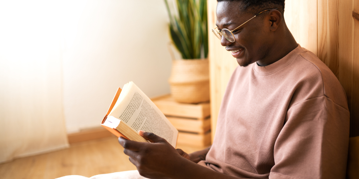 A smiling young man with glasses sits comfortably reading a physical book in a warm, well-lit room with natural wood elements and plants, demonstrating the peaceful benefits of slow reading