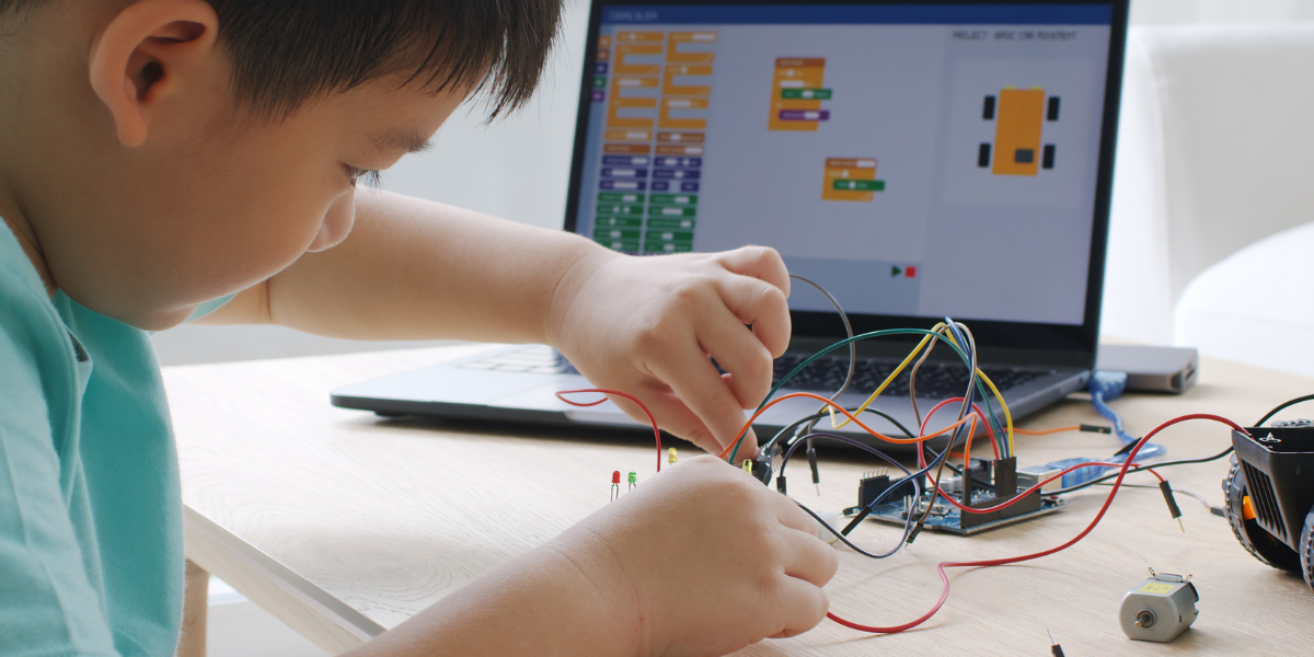 Boy in green shirt working with electronic circuits and coding software on laptop, combining programming skills with hands-on engineering components